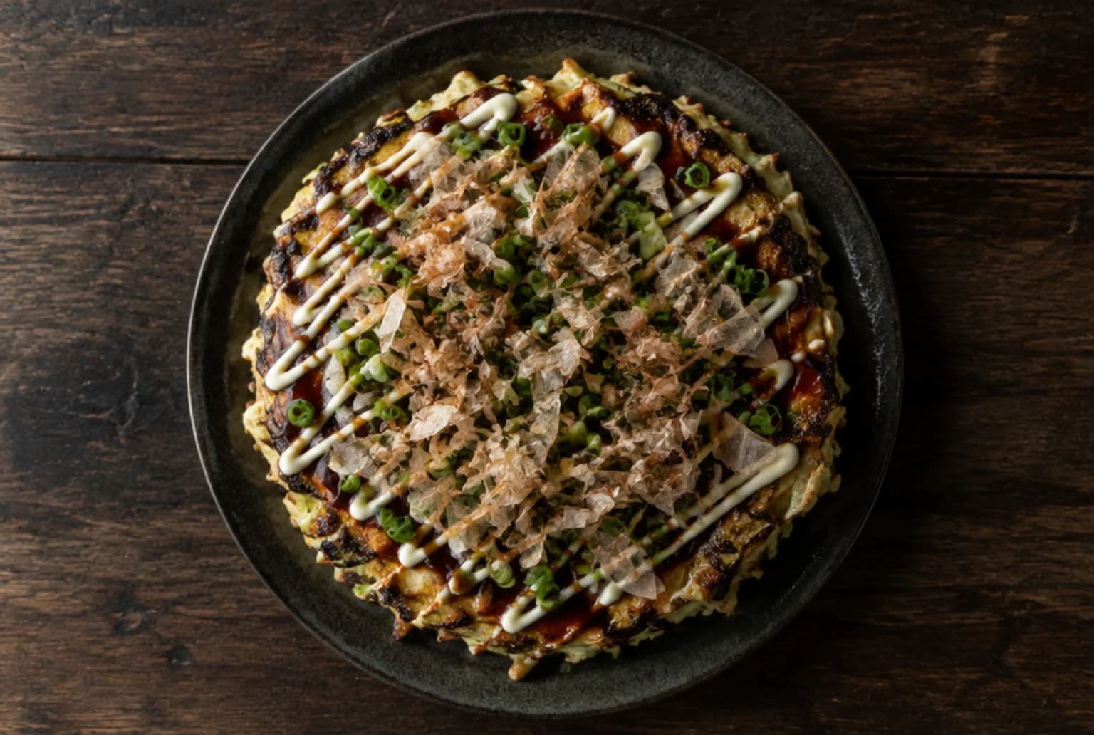Okonomiyaki: runder japanischer Kohlpfannkuchen mit Sauce-Spiralen, Mayonnaise, Bonitoflocken und Frühlingszwiebeln