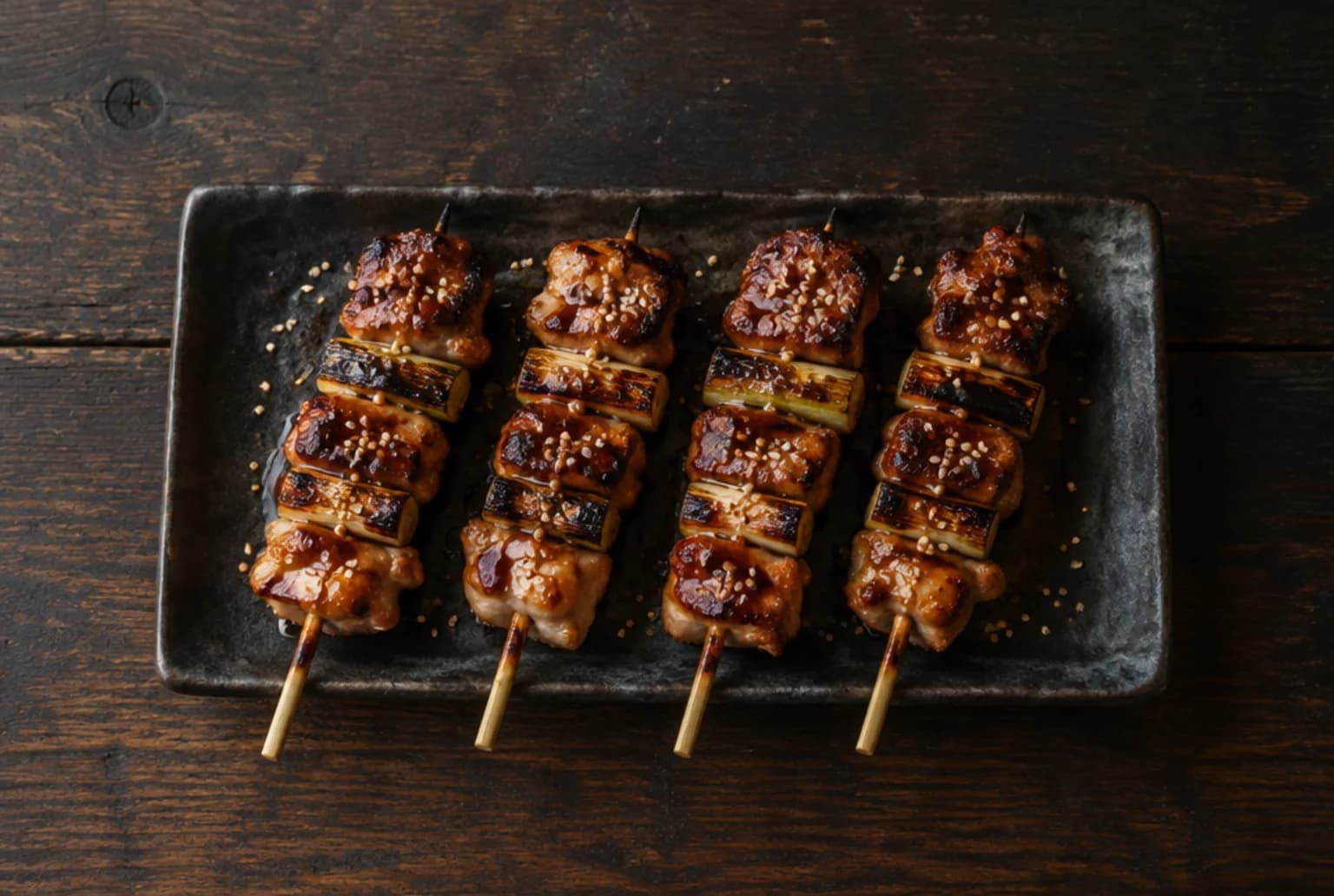 Yakitori: glänzend glasierte Poulet-Spiessli mit Lauch auf dunklem rechteckigem Teller